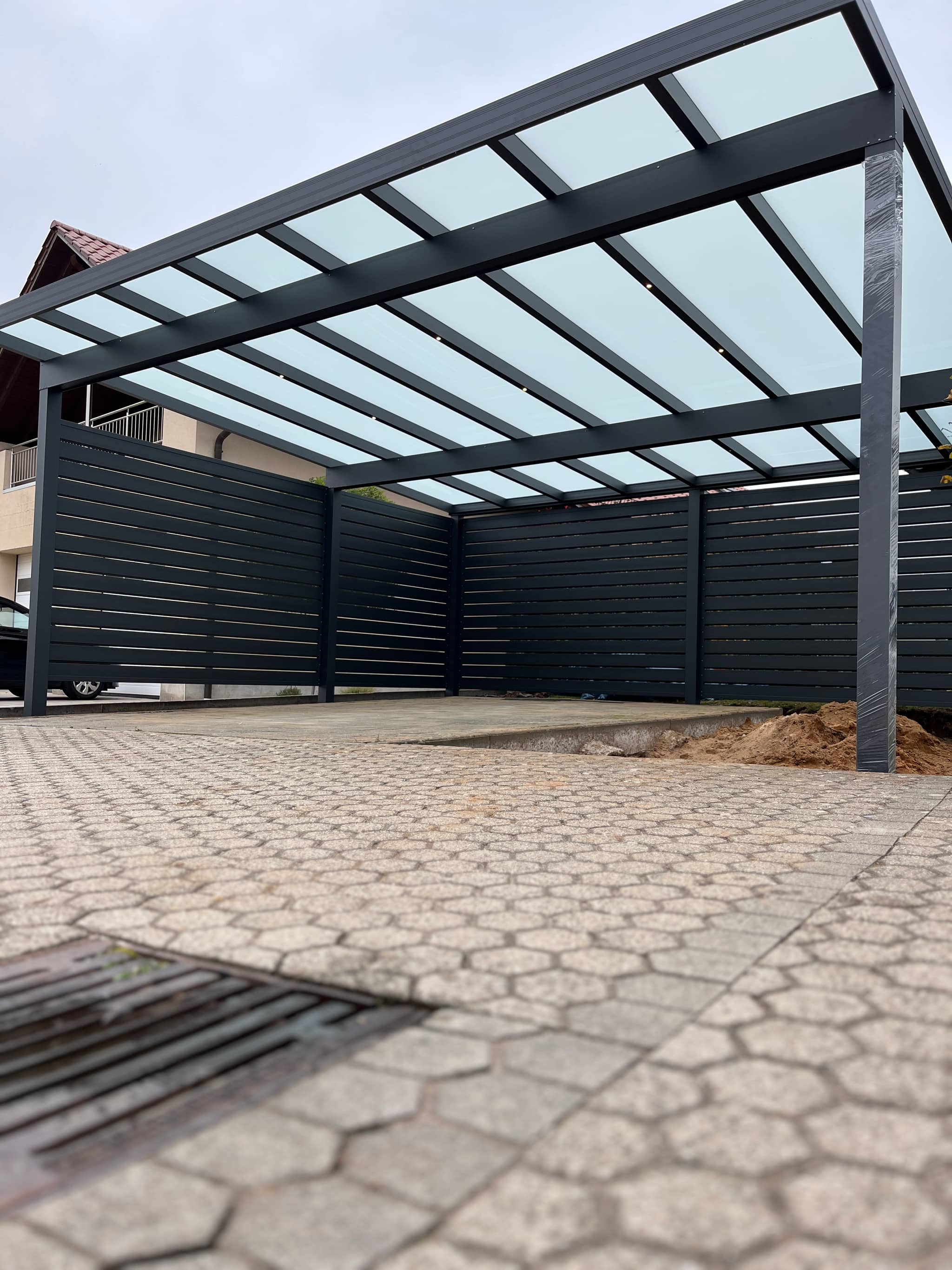 Carport aus Aluminium in Anthrazit mit lichtdurchlässiger Eindeckung und seitlicher Panelwand über gepflasterter Fläche