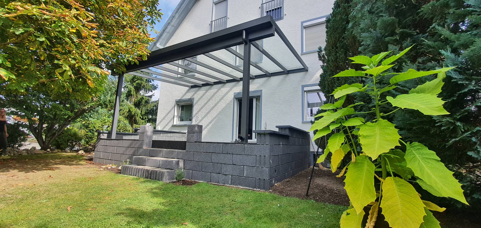 Eingangsüberdachung aus Aluminium in Anthrazit, wandmontiert über einer erhöhten Terrasse mit gemauerter Brüstung