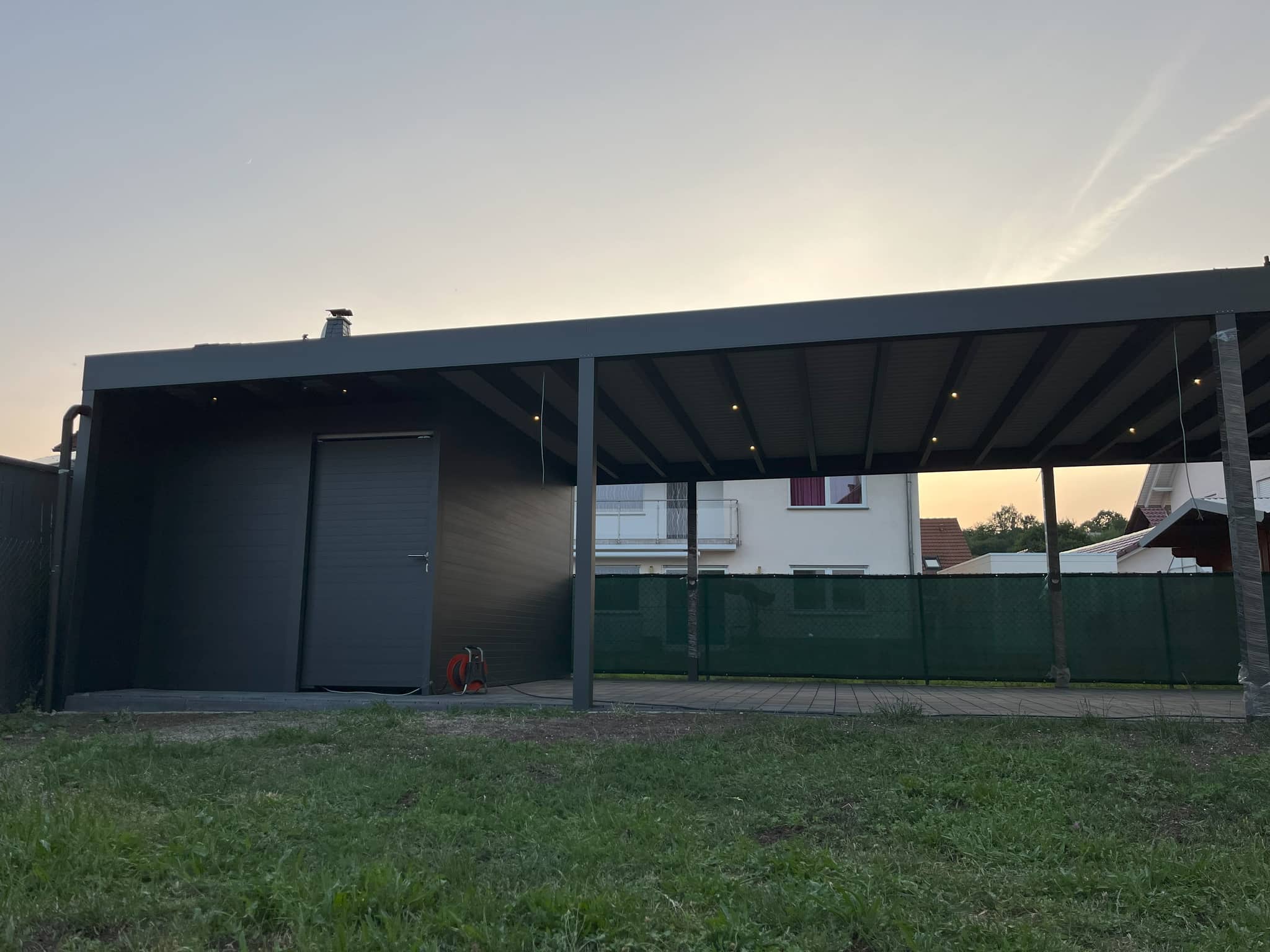 Flachdach-Carport aus Aluminium in Anthrazit mit geschlossenem Abstellraum an der Stirnseite, davor Rasen, Abendhimmel