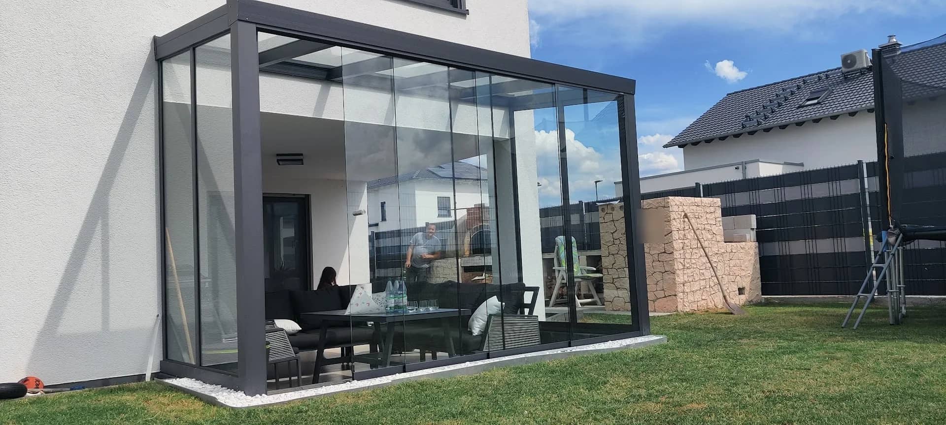 Sommergarten aus Aluminium in Anthrazit mit Glas-Schiebewänden und Lounge-Möbeln an einem weißen Neubau